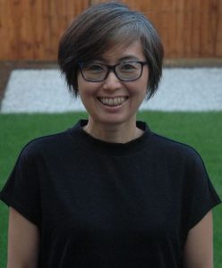 Portrait of Reiko Tanaka. Reiko is standing in her garden. She is smiling and wearing a black top and blue and black glasses.