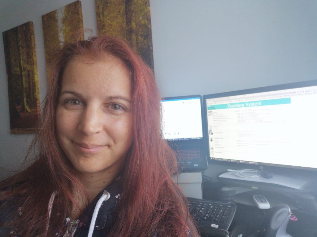 Photo of Edit working from home. Edit is sat in front of two PC monitors. She is smiling and wearing a grey hoodie with white ties. The screen on the right hand side says "Teaching System"