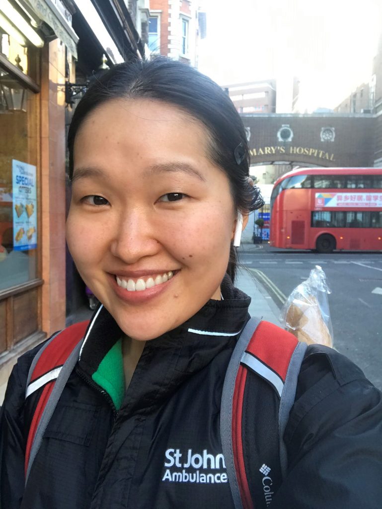 Selfie by Julia Sun. Julia is standing outside smiling, her hair is pulled into a ponytail. She is wearing a black jacket with a green lining and the words "St Johns Ambulance" are embroidered on the left hand side. She is wearing a black and red backpack. There is a red double decker bus in the background