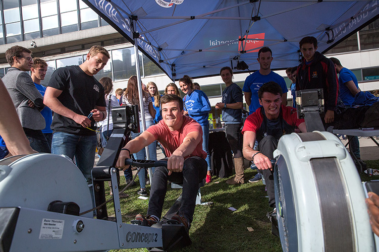 Two men using rowing machines and watched by other students.